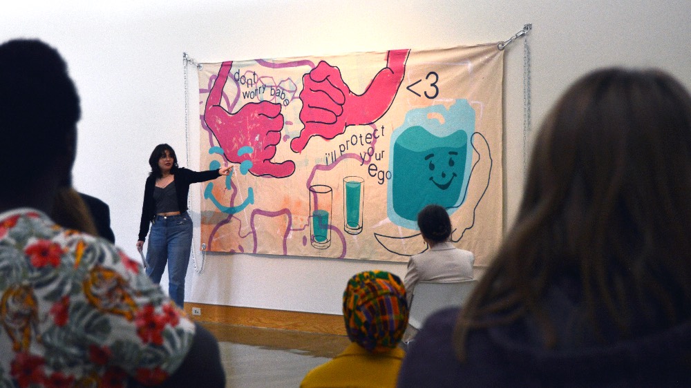 Graduate student points to work hung on the wall while she addresses a crowd